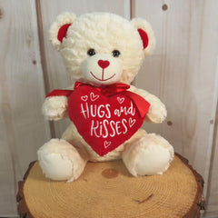 Cream colored bear with a red bow holding a stuffed heart that says, "Hugs and Kisses."