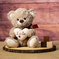 A brown bear that is 10 inches tall while sitting holding a cream Love heart on top of a piece of wood