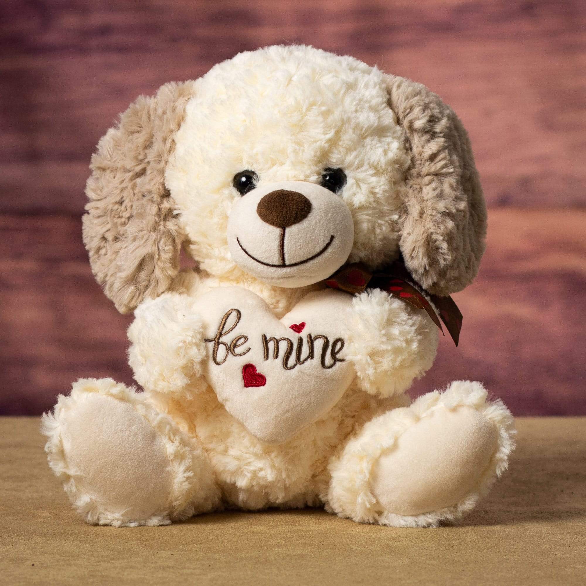 A cream colored dog with brown ears that is 10 inches tall while sitting holding a cream Be Mine heart