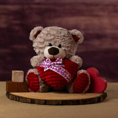 A beige bear that is 10 inches tall while sitting on wooden blocks holding a plush heart wrapped in a Valentine bow tie