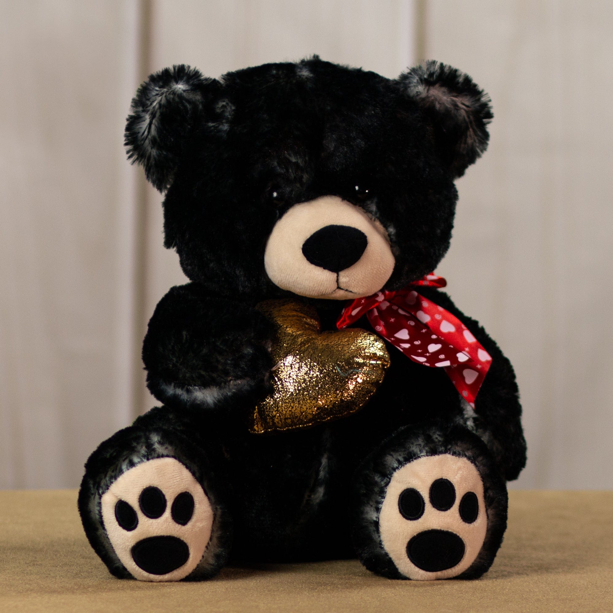 A black bear that is 17 inches tall with paw prints on its feet wearing a red heart covered bow holding a shiny, gold heart