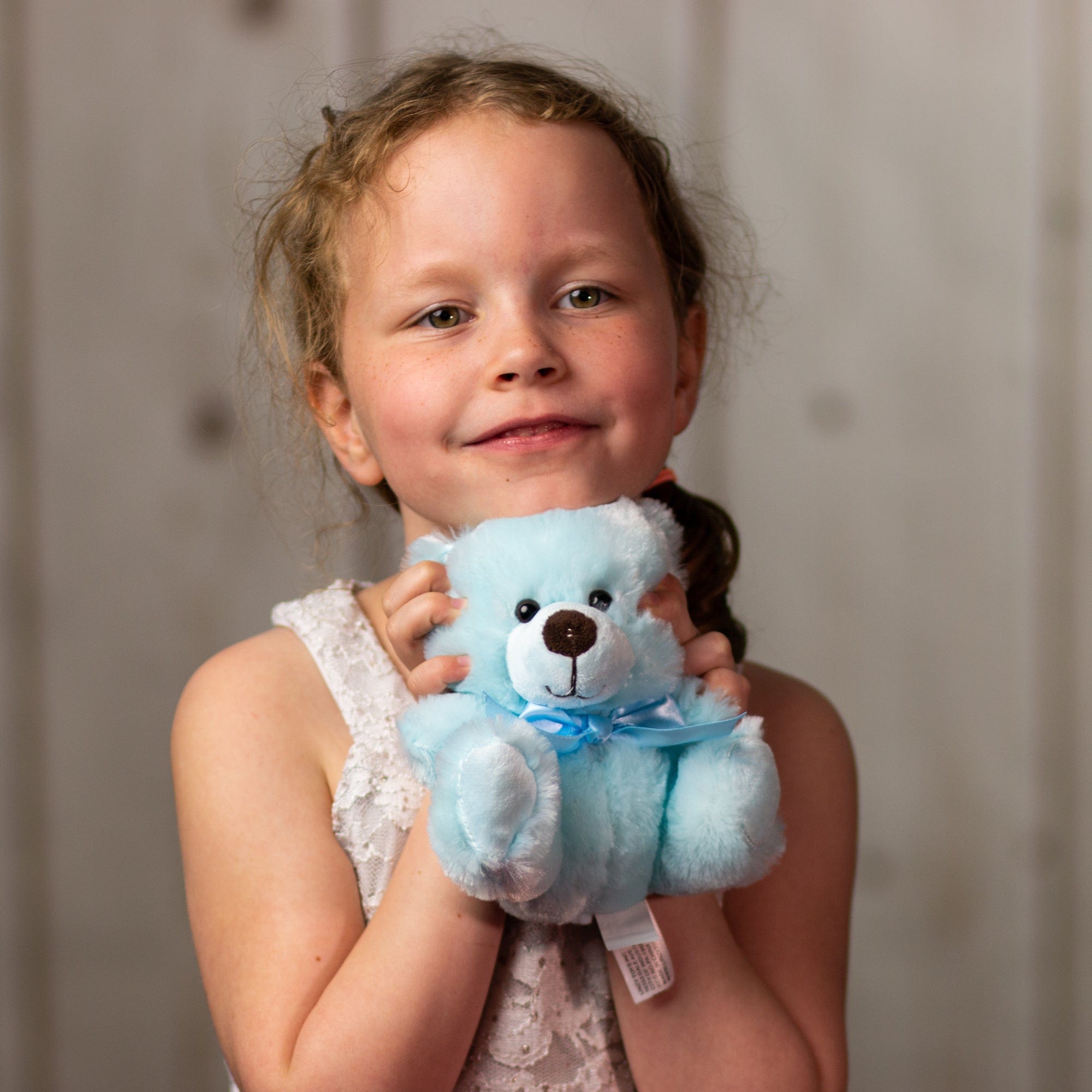 A little girl holding a blue bear that is 6 inches tall while sitting wearing a matching bow