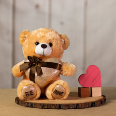 A beige bear that is 11 inches tall while sitting holding a shiny gold gift-wrapped heart with BE MINE embroidered on its feet next to wooden blocks