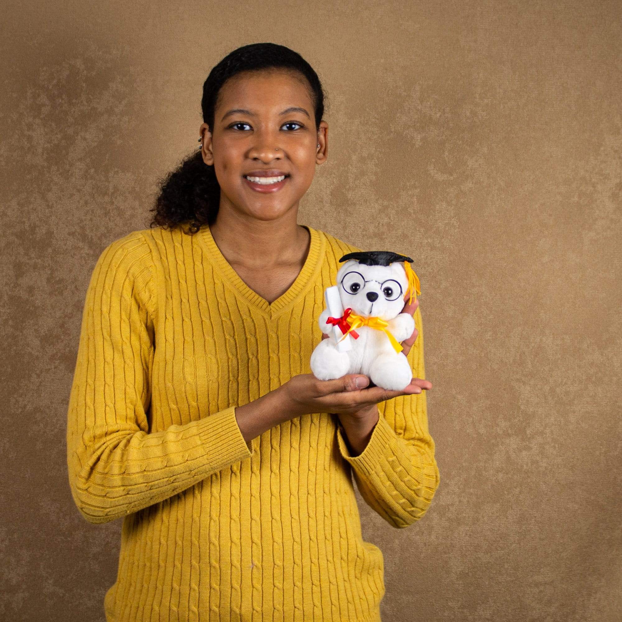 A woman holds a white bear that is 6 inches tall while sitting wearing a graduation hat and glasses and holding a diploma