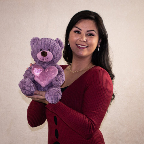 A woman holding a purple bear that is 10 inches tail while sitting holding a heart accented with smaller shiny hearts