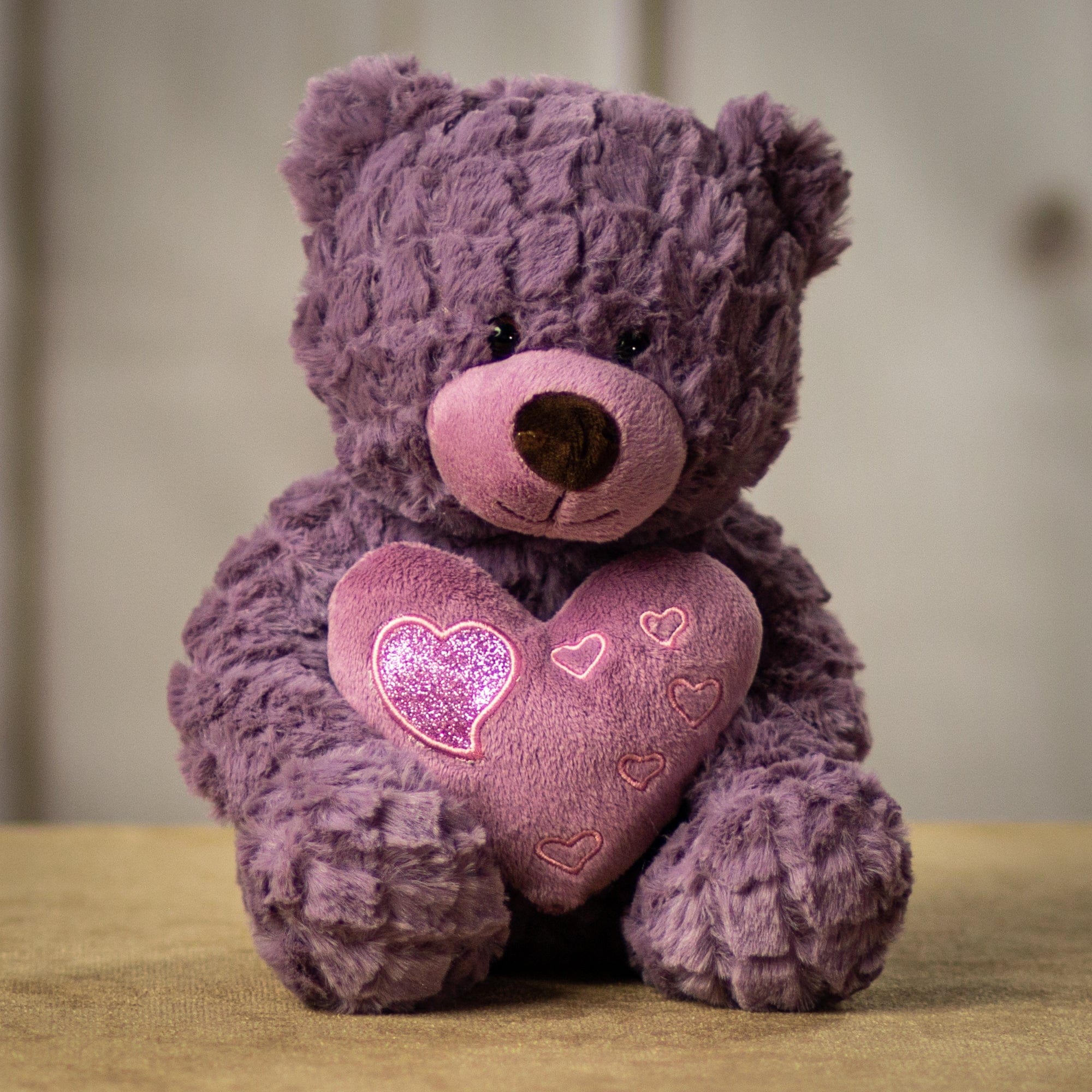 A purple bear that is 10 inches tail while sitting holding a heart accented with smaller shiny hearts