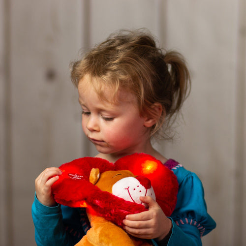 A little girl holding a tan lion that is 11 inches tall while sitting with a red heart wrapped around its head