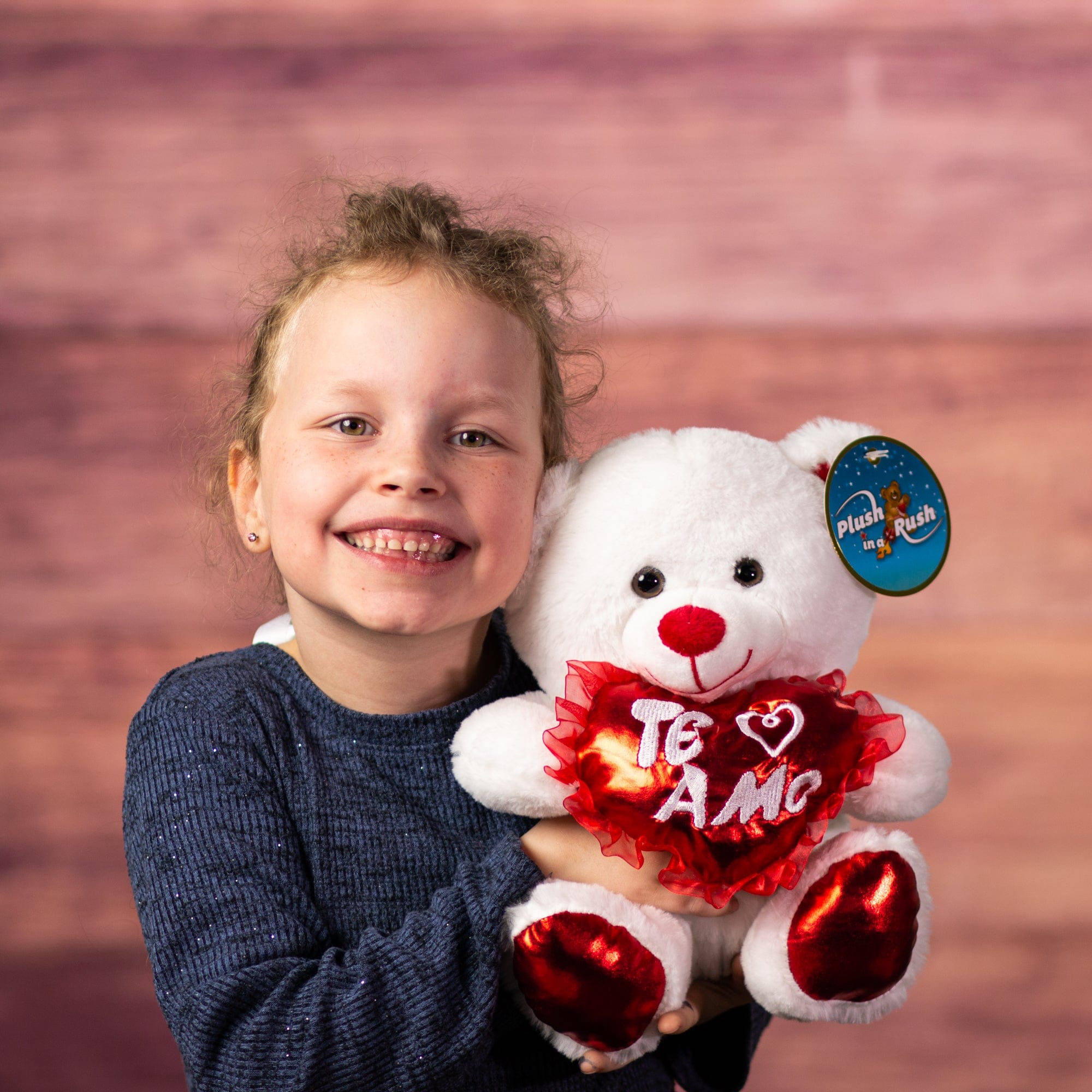 A little girl holding a white bear that is 10 inches tall while sitting with red feet and ears holding a shiny red "Te Amo" heart